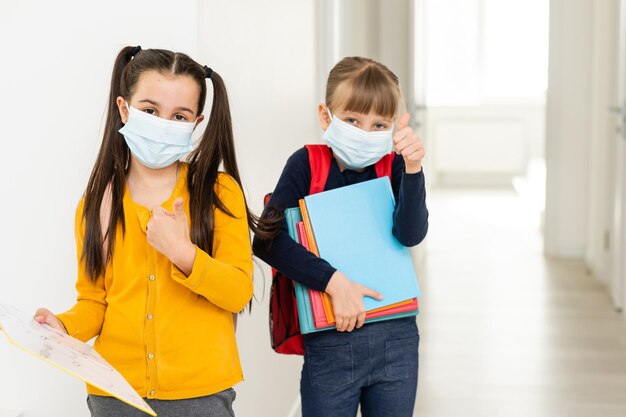 Cute little girls of primary hug each other with the mask on. School girls carry notebooks in hand and greet each other before entering school.