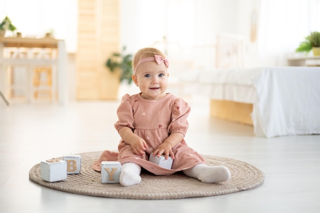 A cute little healthy girl up to a year old in a pink dress made of natural fabric is sitting on a rug in the living room of the house playing with wooden cubes the development of the child at home