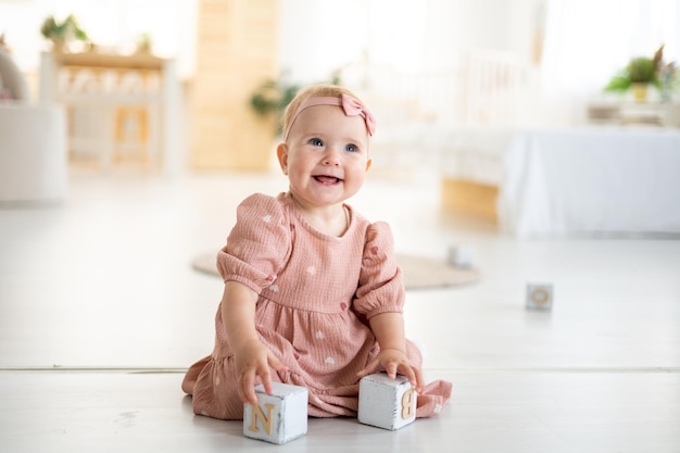 A cute little healthy girl up to a year old in a pink dress made of natural fabric is sitting on a rug in the living room of the house playing with wooden cubes the development of the child at home