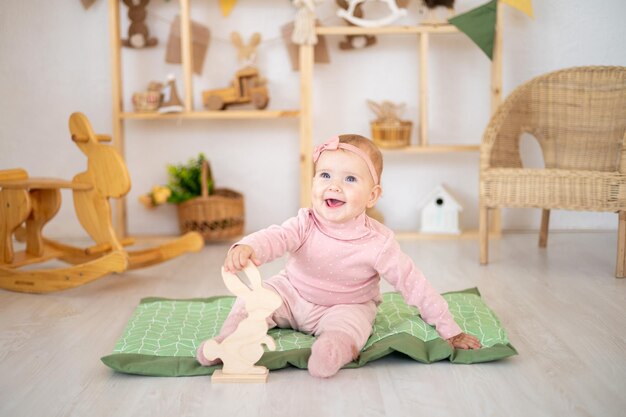 A cute little healthy girl up to a year old in a pink suit made of natural fabric is sitting on a rug in a children's room with wooden educational toys looking at the camera smiling