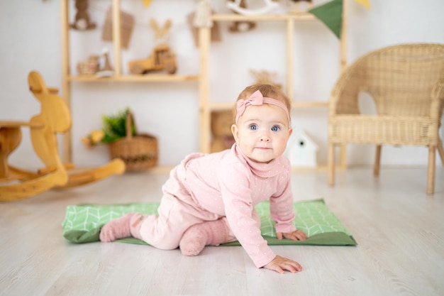 A cute little healthy girl up to a year old in a pink suit made of natural fabric is sitting on a rug in a children's room with wooden educational toys looking at the camera smiling