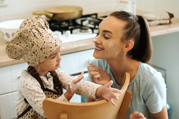 Cute little homecook girl with her beautiful mother makes pancakes in white kitchen