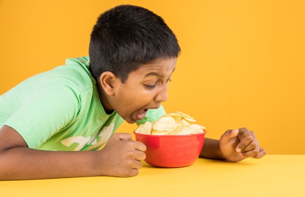 Cute little indian or asian boy eating chips or potato wafers in big red bowl, over yellow background