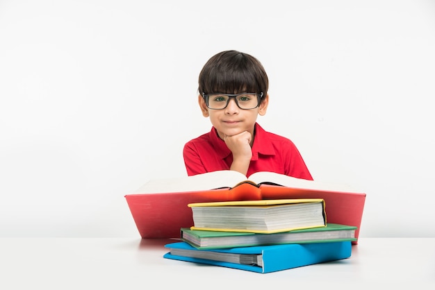 Simpatico ragazzino indiano o asiatico che tiene o legge un libro sul tavolo da studio o sul pavimento bianco, isolato su sfondo bianco