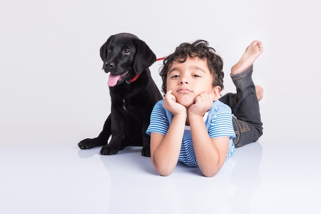 Cute little Indian or Asian boy playing with Black Labrador Retriever puppy while lying or sitting isolated on white background