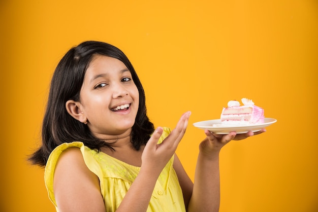 Cute little Indian or Asian girl child eating piece of Strawberry or Chocolate flavoured pastry or cake in a plate. Isolated over colourful background