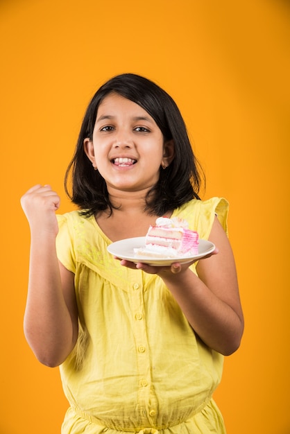 Cute little Indian or Asian girl child eating piece of Strawberry or Chocolate flavoured pastry or cake in a plate. Isolated over colourful background