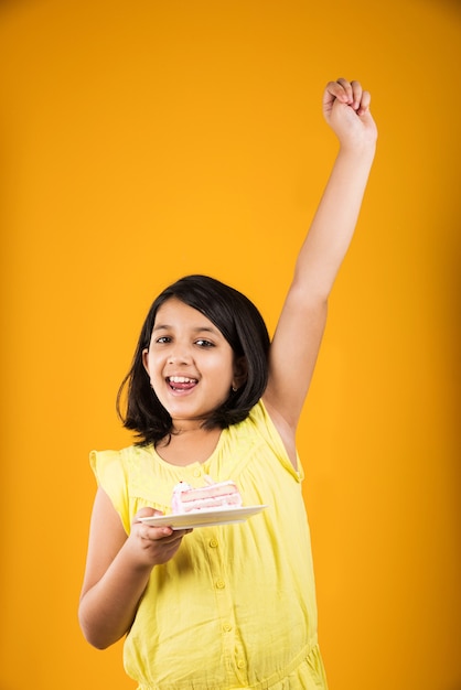 Cute little Indian or Asian girl child eating piece of Strawberry or Chocolate flavoured pastry or cake in a plate. Isolated over colourful background