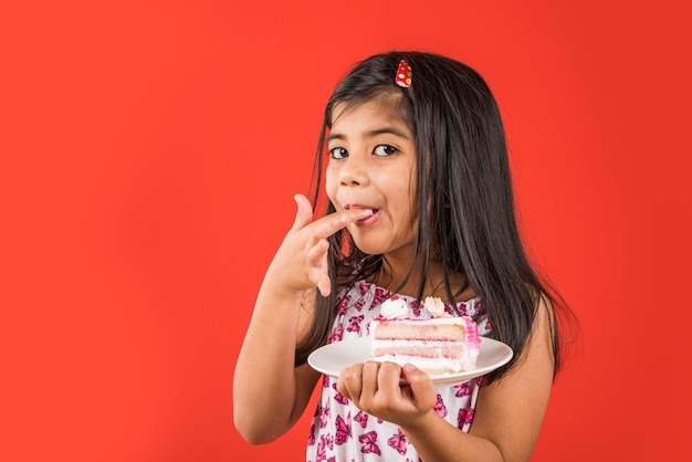 Милый маленький индийский или азиатский ребенок девочка ест кусок теста или пирога со вкусом клубники или шоколада в тарелке. Изолированные на красочном фоне