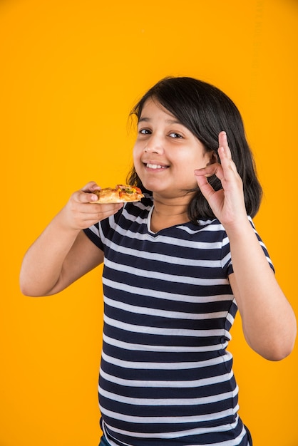 Cute little Indian or Asian girl child eating tasty Burger, Sandwich or Pizza in a plate or box. Standing isolated over blue or yellow background.