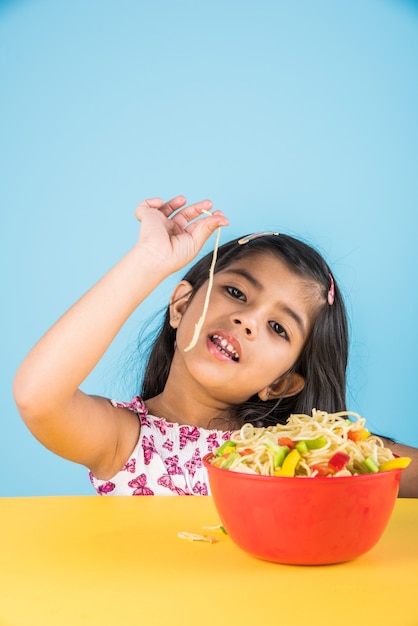 カラフルな背景の上に分離されたフォークや箸でおいしい中華麺を食べるかわいい小さなインドやアジアの女の子の子供