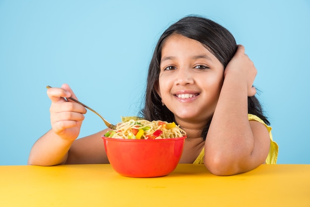 Cute little Indian or Asian girl child eating yummy Chinese Noodles with fork or chopsticks, isolated over colourful background