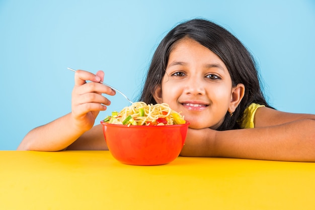 Милая маленькая индийская или азиатская девочка ест вкусную китайскую лапшу вилкой или палочками для еды, изолированные на красочном фоне