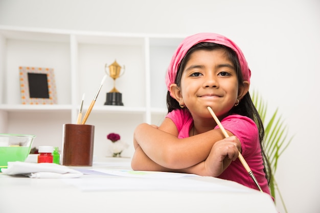 Симпатичная маленькая индийская или азиатская девочка, которая любит рисовать или рисовать кистью и раскрашивать бумагу дома