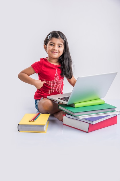 Cute little Indian or Asian girl child studying on laptop or working on school project while lying or sitting on the floor, isolated over white background