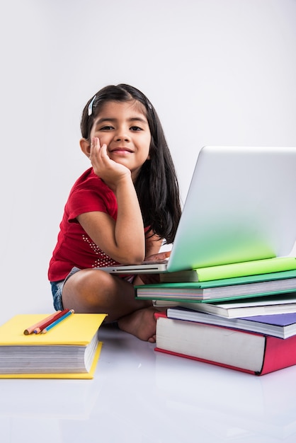 Cute little Indian or Asian girl child studying on laptop or working on school project while lying or sitting on the floor, isolated over white background