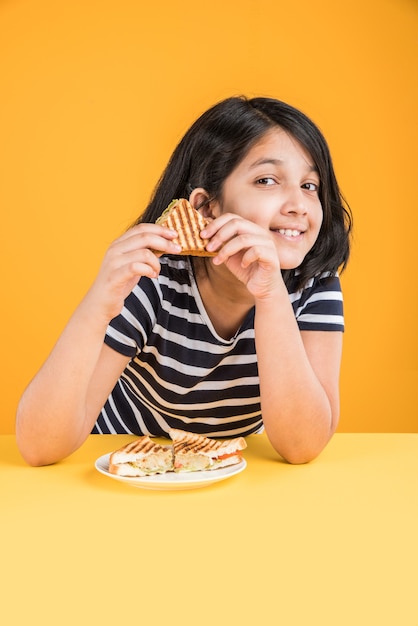 Carina bambina indiana o asiatica che mangia un gustoso hamburger, panino o pizza in un piatto o in una scatola. in piedi isolato su sfondo blu o giallo.