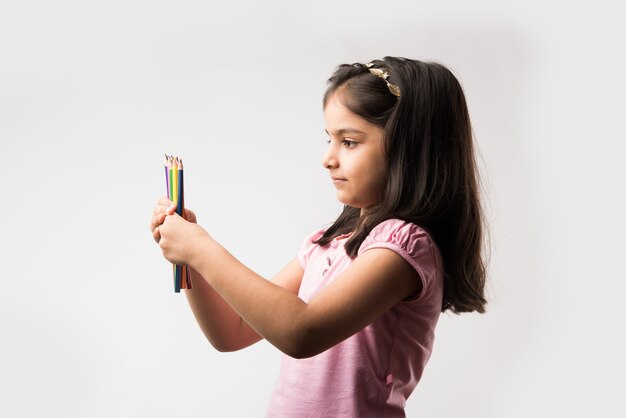 Cute little Indian or asian girl holding coloured pencils, standing isolated over white background