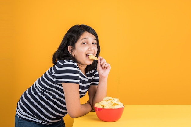 Cute little indian or asian girl kid eating chips or potato wafers in big red bowl, over yellow background