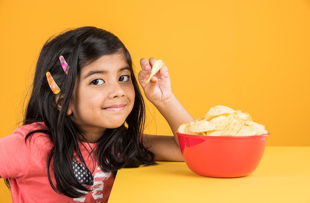 Cute little indian or asian girl kid eating chips or potato wafers in big red bowl, over yellow background