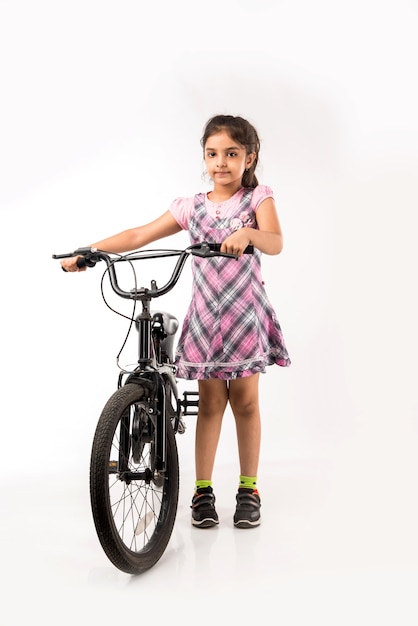 Милая маленькая индийская или азиатская девушка катается на велосипеде, изолированном на белом фоне, держа воздушные шары
