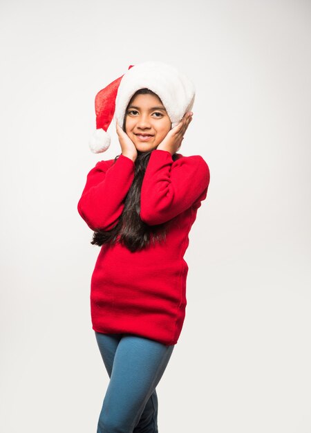 Cute Little Indian Asian Girl wearing Santa Hat celebrating christmas while standing isolated over plain background