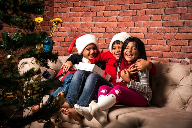 Cute little Indian Asian kids celebrating Christmas at home with Santa Hat, Gifts and Xmas Tree