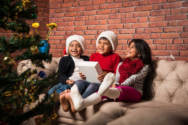 Cute little Indian Asian kids celebrating Christmas at home with Santa Hat, Gifts and Xmas Tree