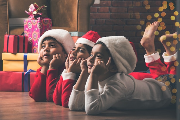 Photo cute little indian asian kids celebrating christmas at home with santa hat, gifts and xmas tree