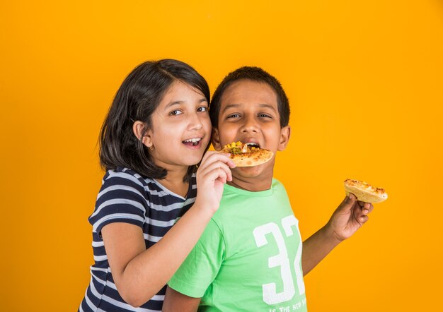 プレートまたはボックスでおいしいハンバーガー、サンドイッチまたはピザを食べるかわいい小さなインドまたはアジアの子供たち。青または黄色の背景の上に孤立して立っています。