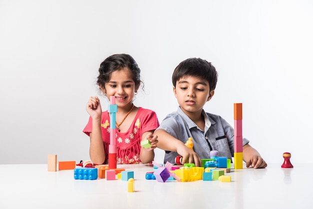 Cute little Indian asian kids playing with colourful plastic toys or blocks while sitting at table or isolated over white background