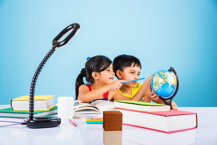 फोटो cute little indian or asian kids studying on study table with pile of books, educational globe, isolated over light blue colour
