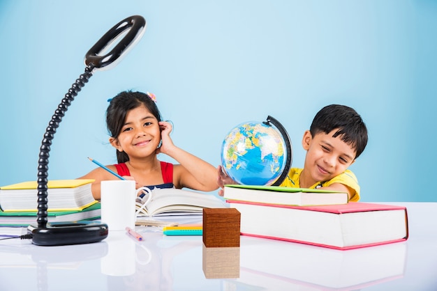 Cute little indian or asian kids studying on study table with\
pile of books, educational globe, isolated over light blue\
colour