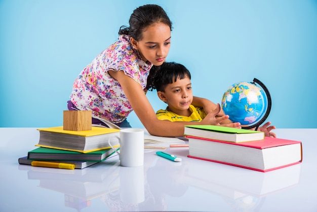 Cute little indian or asian kids studying on study table with\
pile of books, educational globe, isolated over light blue\
colour