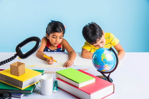 水色で隔離の本、教育グローブの山で勉強のテーブルで勉強しているかわいい小さなインドやアジアの子供たち