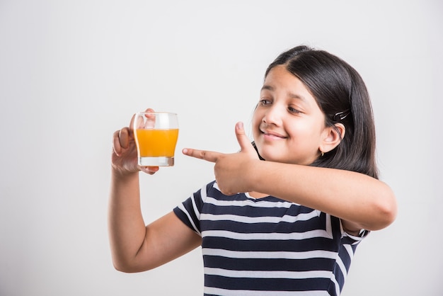 Cute little Indian or Asian playful girl drinking fresh mango or orange juice or cold drink or beverage in a glass, isolated over  white background