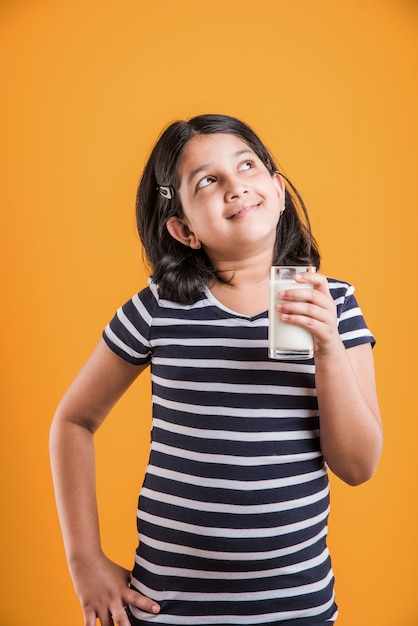 Милая маленькая индийская или азиатская игривая девочка держит или пьет стакан, полный молока, изолированные на красочном фоне