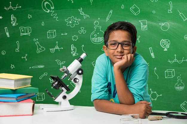 Cute little Indian Asian School Boy student experimenting or Studying Science in Laboratory, Over green chalkboard background with educational doodles