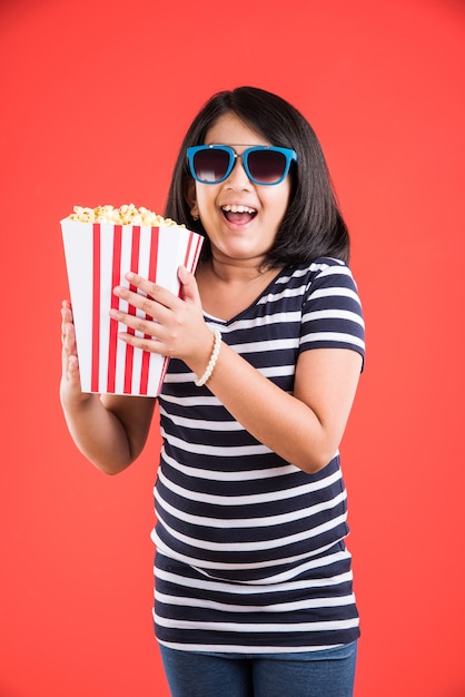 Carina bambina indiana che mangia popcorn mentre indossa occhiali da sole o occhiali 3d in un teatro. in piedi isolato su sfondo colorato
