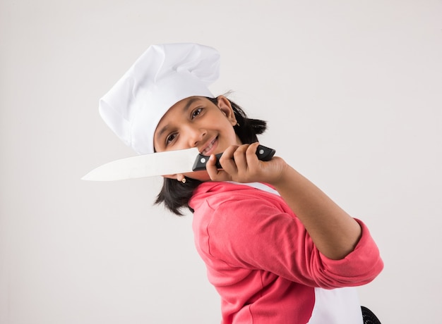 Cute little Indian kid or girl dressed as a Chef and holding cooking utensils or vegetables, standing isolated over white background