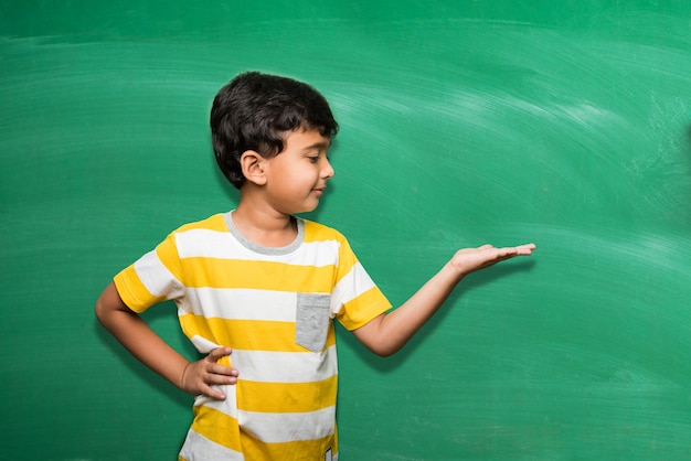 Cute little Indian school kid boy in hand stretched pose over green chalkboard or chalk board background holding books, victory cup etc, isolated