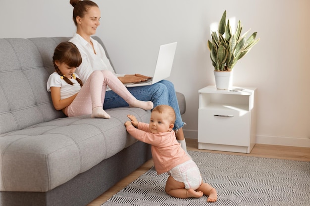 Cute little infant baby girl crawling to her mother sitting on sofa with her elder daughter and working online on laptop, female freelancer earning money while having maternity leave.