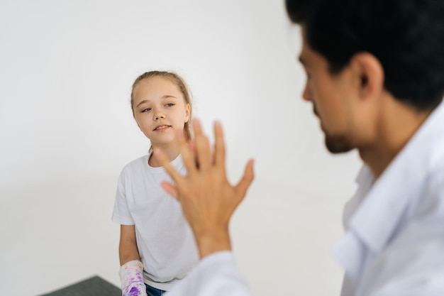 Cute little injured girl with broken hand wrapped in plaster\
bandage giving high five to friendly male doctor at checkup meeting\
on white isolated background
