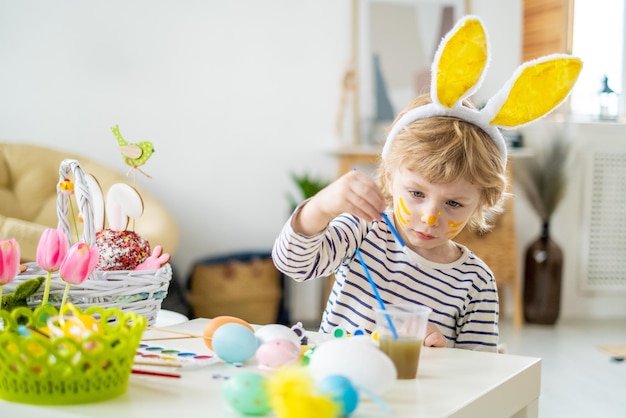 Ragazzino carino con la fascia per le orecchie da coniglio e il viso compongono le uova da colorare per le vacanze di pasqua con vernice e pennello che decorano le uova sviluppando creatività e immaginazione trascorrendo felicemente il tempo a casa