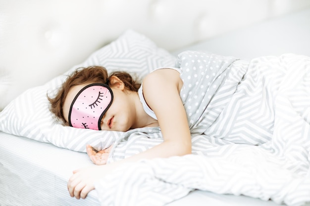 Foto piccola ragazzina carina con una maschera da notte rosa che dorme sul letto