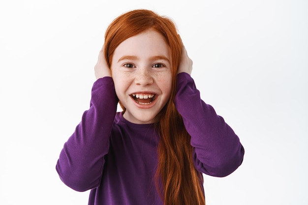 Photo cute little kid with red hair shut her ears and smiling, acting childish, unwilling to listen, misbehave, laughing and having fun, standing over white background