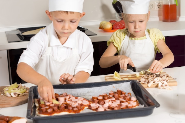 Симпатичные маленькие дети в одежде шеф-повара, делая вкусную пиццу на кухне.