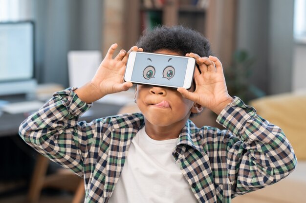 Cute little mixed-race boy in white t-shirt and checkered shirt holding smartphone with funny eyes on screen in front of his face