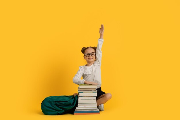 Cute little nerdy schoolgirl sitting with pile of books and raising hand