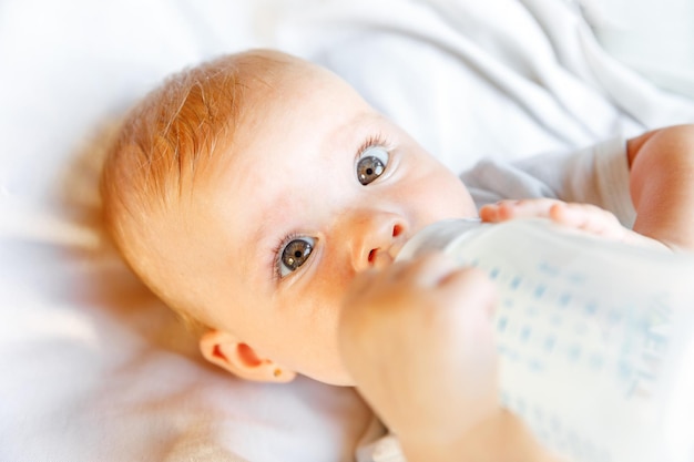 Cute little newborn girl drinking milk from bottle and looking at camera on white background infant ...
